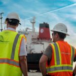 Two men standing in front of a heavy lift vessel that is unloading cranes. The taller man, on the left, is wearing a yellow high-visibility vest with the UTC Overseas logo on the back. The second man, on the right, is wearing an orange high-visibility vest with the Konecranes logo on the back. Both are wearing white hard hats.