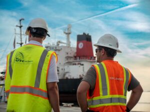 Two men standing in front of a heavy lift vessel that is unloading cranes. The taller man, on the left, is wearing a yellow high-visibility vest with the UTC Overseas logo on the back. The second man, on the right, is wearing an orange high-visibility vest with the Konecranes logo on the back. Both are wearing white hard hats.