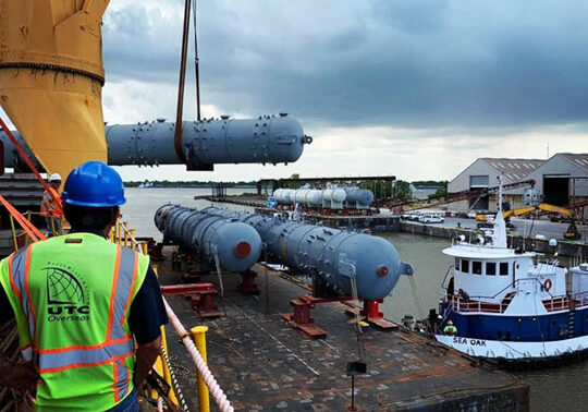 L-R: man wearing light blue construction hat and reflective vest with UTC Overseas logo on back. Man standing on heavy-lift vessel, yellow crane in background is lifting one of three large pieces of oil & gas equipment. To the man's right, two pieces are already transloaded to the barge. Bright white and blue tug is on the right, keeping barge in place next to vessel. Cloudy skies above.