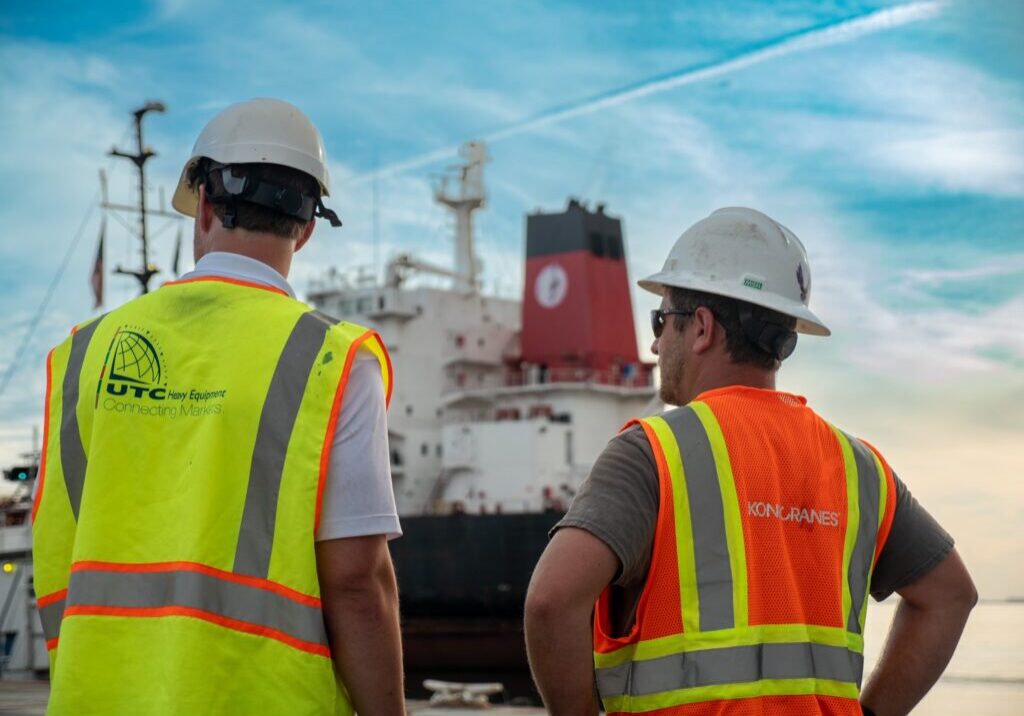 Two men standing in front of a heavy lift vessel that is unloading cranes. The taller man, on the left, is wearing a yellow high-visibility vest with the UTC Overseas logo on the back. The second man, on the right, is wearing an orange high-visibility vest with the Konecranes logo on the back. Both are wearing white hard hats.